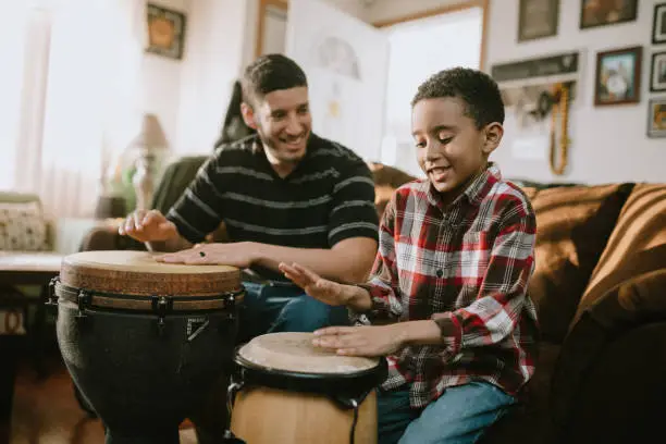 A dad plays djembe drum with his son in their living room, teaching the boy technique on how to play the instrument.