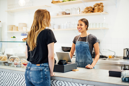 Smiling young woman barista talking with a female customer while standing behind the counter of a trendy cafe. Waitress taking order from customer in coffee shop.