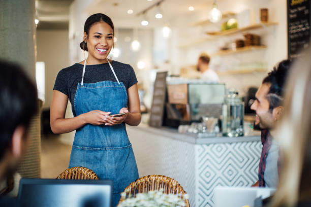 Beautiful waitress  at cafe taking order from customers Beautiful female waiter at cafe with mobile phone taking order from customers sitting at table. Young waitress in apron holding a smart phone to take order. waitress stock pictures, royalty-free photos & images