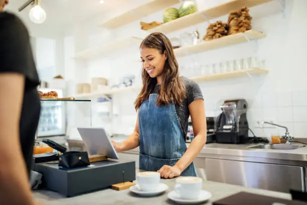 Female barista standing at cafe counter entering customer order on digital tablet. Cafe owner taking order from customer at coffee shop counter.