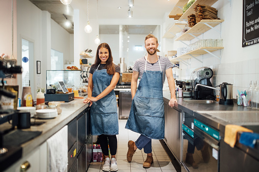 Two young multiracial baristas in aprons standing in kitchen of coffee shop. Smiling man and woman standing by kitchen counter of cafe and looking at camera.