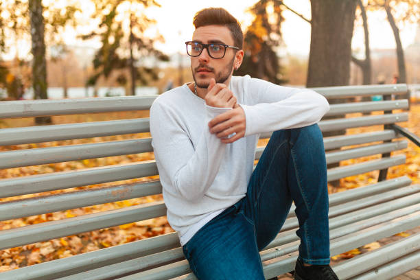 istock Young handsome hipster guy is sitting in autumn park on a bench. Bored alone. A student walks in the park. 1060919836