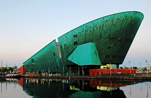 Amazing wide angle view of the Nemo Science Museum of Amsterdam reflected in the water at twilight. This modern building with a futuristic copper green ship-like design was opened in 1997. It stands on top of the IJ-tunnel.  The roof of the building is a large and very sunny terrace which in summer has been turned into an entertaining city beach.
