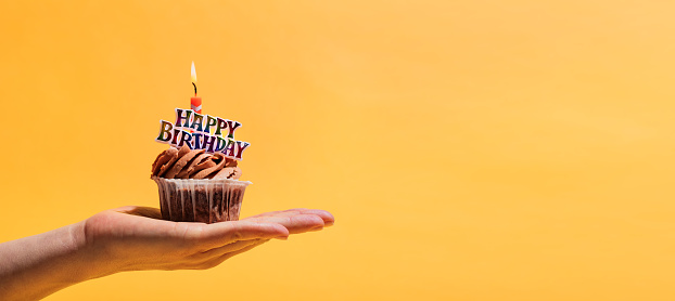 Woman's hand holding decorated birthday muffin against yellow background. Birthday party and celebration.
