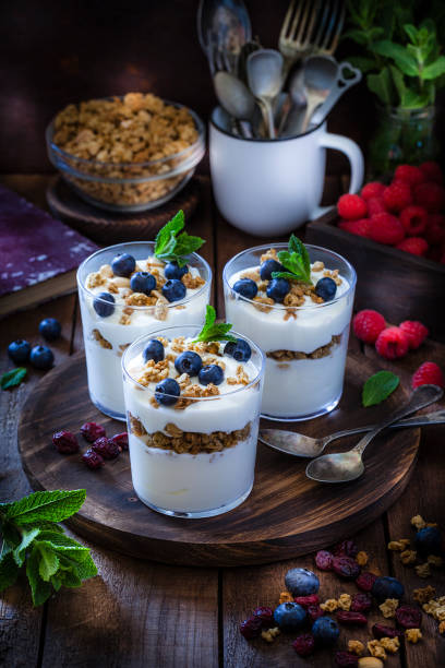 Three homemade yogurt with berries and granola served in glass containers shot on rustic wooden table. The containers are arranged in a round wooden tray and are surrounded by berry fruits and granola. Low key DSRL studio photo taken with Canon EOS 5D Mk II and Canon EF 100mm f/2.8L Macro IS USM.