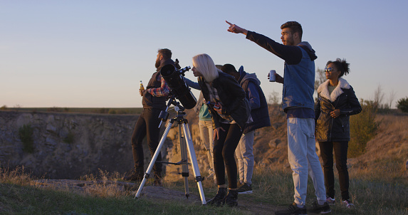 Wide shot of happy adult friends stargazing together and using a professional telescope and pointing at the sky