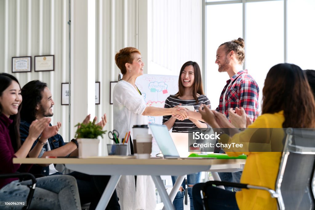 Atraente madura mulher asiática, apertando a mão com hipster jovem barba branca com bem-vindo novo empregado, felicitando bem sucedido ou feliz com o negócio lidar enquanto suporte aplaudir a equipe juntos. - Foto de stock de Funcionário royalty-free