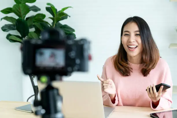 Photo of Young attractive asian woman blogger or vlogger looking at camera and talking on video shooting with technology. Social media influencer people or content maker concept in relax casual style at home.