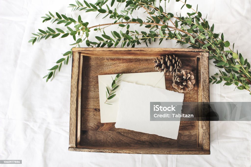 Christmas or winter wedding mock-up scene. Blank cotton paper greeting cards, old wooden tray, pine cones and green Eucalyptus parvifolia branch.White bed linen background. Flat lay, top view. Christmas or winter wedding mock-up scene. Blank cotton paper greeting cards, old wooden tray, pine cones and green Eucalyptus parvifolia branch, white bed linen background. Flat lay, top view. Wedding Stock Photo