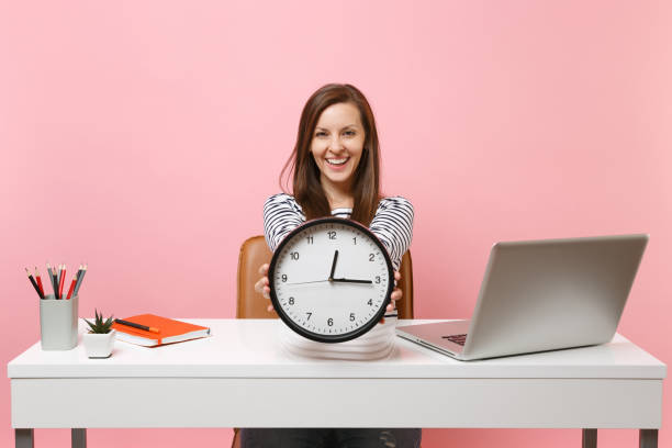 istock Smiling woman holding round alarm clock while sit and working on project at white desk with contemporary pc laptop isolated on pastel pink background. Achievement business career concept. Copy space. 1060990622