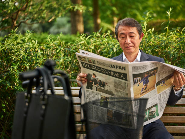 A mature Japanese man in a business suit, reading a newspaper in an outdoor setting in Tokyo, Japan.