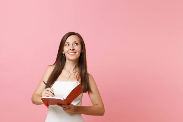 istock Dreamy bride woman in wedding dress looking up write note in notebook planning wedding choosing of staff wedding organizers isolated on pink pastel background. Wedding to do list. Celebration concept. 1060994746