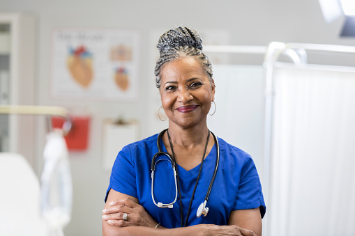 In this portrait, an African American senior female doctor smiles for the camera in an exam room with her arms folded.  She is wearing scrubs and a stethoscope.