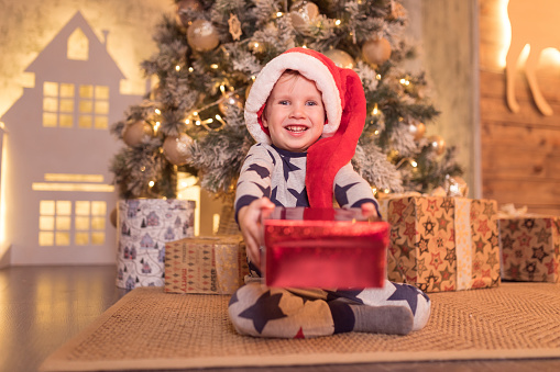 Little kid boy in pajamas with stars and in red Santa hat is sitting under the Christmas tree
