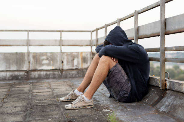 Depression problems Depressed, desperate young man sitting on the building rooftop terrace, wearing hoodie, holding head in hands self destructive stock pictures, royalty-free photos & images