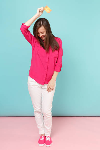 Full length portrait of smiling young woman in rose shirt blouse white pants hold credit card isolated on bright pink blue pastel wall background studio. Fashion lifestyle concept. Mock up copy space retrato de longitud completa de la sonriente mujer joven en camisa rosa blusa, pantalones blancos mantenga aislado en el estudio de fondo de pared pastel azul rosado brillante de tarjeta de crédito. concepto de estilo de vida de moda. simulacros de espaci