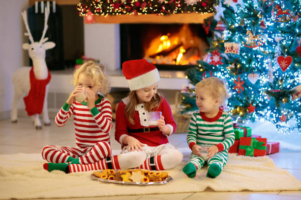 Children at Christmas tree and fireplace drink hot chocolate and eat cookies on Xmas eve. Family with kids celebrating Christmas at home. Boy and girl opening presents. Holiday gifts for kid.