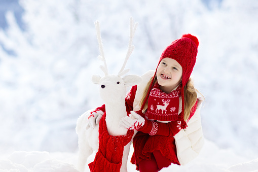 Child playing with reindeer in snow on Christmas vacation. Winter outdoor fun. Kids play in snowy park on Xmas eve. Little girl in knitted sweater, hat, scarf and mittens with Christmas decoration.