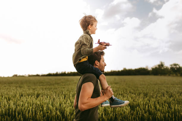Photo of a cheerful little boy, being supported to be a pilot, by his father who is holding him on shoulders