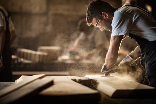 Young carpenter using sander while working on a piece of wood. Manual worker using sander while working on a wood in carpentry workshop. blue collar worker stock pictures, royalty-free photos & images