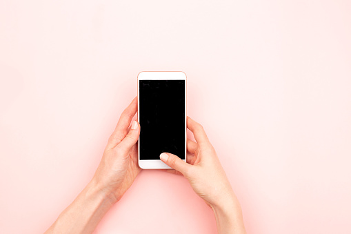 Woman's hands are using a smartphone. Pastel pink background. Mock up for your design. Copy space. Flat-lay, top view.
