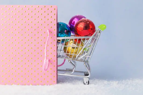 Christmas shopping concept. Shopping cart, pink bag, christmas multi-colored balls, on a light background. Copy space