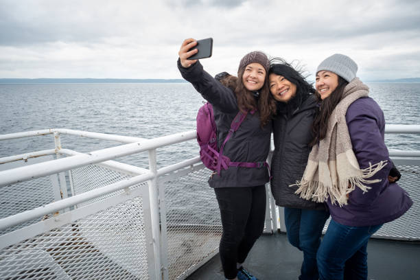 Winter Travel on Ferry, Mother and Teen Daughters Taking Selfie Asian mother and Eurasian teen daughters on deck of ferry between Vancouver and Nanaimo, British Columbia, Canada. ferry stock pictures, royalty-free photos & images