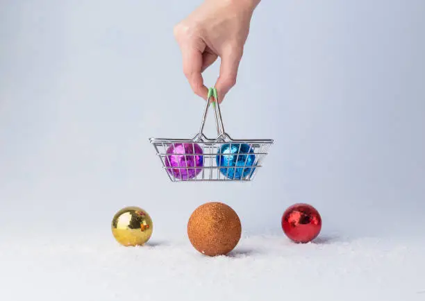 Male hand holds shopping basket. Christmas multi-colored balls, on light background. Copy space. Christmas shopping minimal concept.