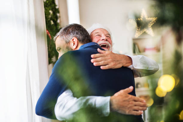 un padre senior con un sombrero de santa y el hijo adulto de pie junto a la ventana, abrazos. - christmas window santa claus lighting equipment fotografías e imágenes de stock