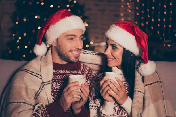 Noel christmastime concept. Profile side view photo of two lovely, careless, carefree persons sit on cozy couch or sofa in living room hold mulled-wine drinks look to each other with beaming smile