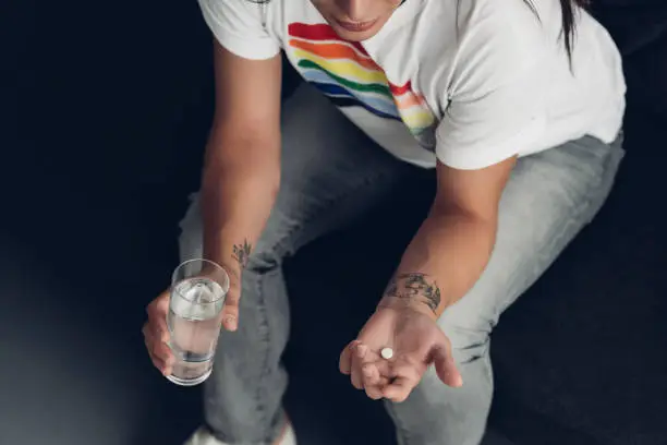 cropped shot of young transgender man with pill and glass of water sitting on couch