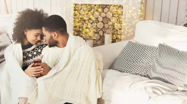 Loving african-american couple embracing, covered with plaid and drinking mulled-wine in cozy light room