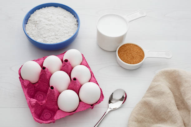 The image shows a carton of fresh brown eggs placed on a kitchen surface along with a small bowl of butter and a pile of flour, indicating a setup for baking or cooking