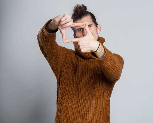 istock Young man focusing with fingers, framing with hands on grey background, copy space. Close up portrait man in warm knitted sweater holding his hands out in a framing gesture. 1061291062