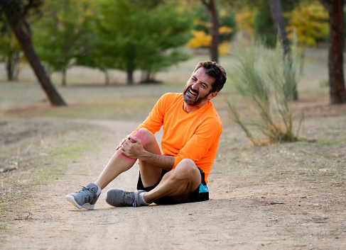 Young fit man holding knee with his hands in pain after suffering muscle injury broken bone leg pain sprain or cramp during a running workout in park outdoors in sport training running injury.