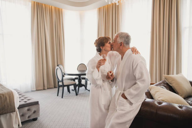 Happy Anniversary Celebration in Hotel Mature couple are kissing in their hotel room with a glass of champagne. They are both wearing dressing gowns. kissing on the mouth stock pictures, royalty-free photos & images