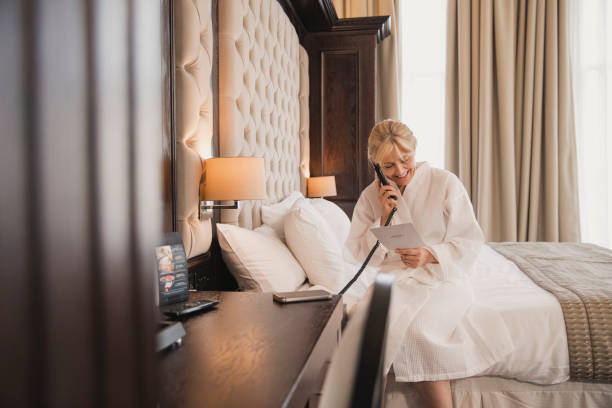 Ordering Room Service Mature woman is sitting on her bed in her hotel room, using the phone to order room service. room service stock pictures, royalty-free photos & images