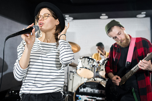 Waist up portrait of contemporary music band rehearsing in studio, focus on young woman singing in foreground, copy space