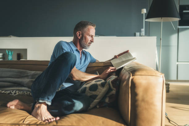 uomo barbuto midaged libro di lettura sul divano - reading book men sofa foto e immagini stock