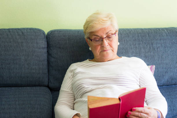 istock Senior woman with eyeglasses sitting on sofa reading red cover book 1061420410