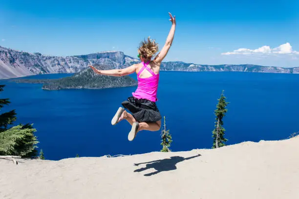 Adult female girl wearing bright clothing jumps at Crater Lake National Park in Oregon. Concept for freedom, confidence, solo female travel