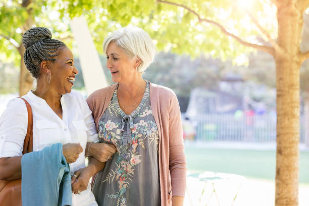 Senior female friends link arms and stroll through the park Two beautiful senior women are linking arms and strolling through an outdoor park together. locking-arms stock pictures, royalty-free photos & images