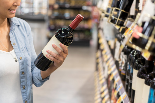 While shopping alone in a supermarket, a mid-adult woman reads the label on a bottle of red wine in the beverage aisle.