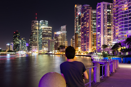 Back view of unrecognizable guy leaning on railing on city embankment and looking at illuminated skyscrapers in Brisbane, Australia