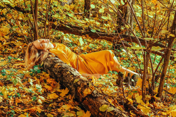 giovane bella ragazza con un lungo abito giallo sdraiato a terra del parco autunnale con foglie gialle cadute - bench sitting tree apple foto e immagini stock