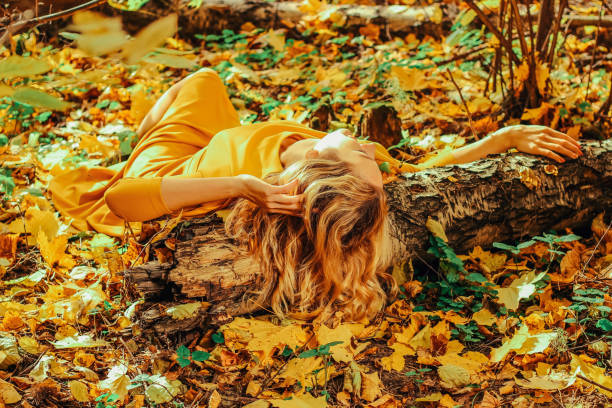 giovane bella ragazza con un lungo abito giallo sdraiato a terra del parco autunnale con foglie gialle cadute - bench sitting tree apple foto e immagini stock