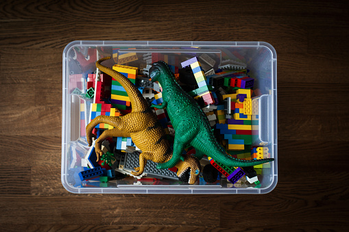 top view closeup of transparent toy container box with plastic T Rex dinosaurs and colored construction blocks on dark brown wooden parquet floor background