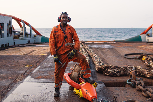 Seaman AB or Bosun on deck of offshore vessel or ship , wearing PPE personal protective equipment - helmet, coverall, lifejacket, goggles. He holds hammer