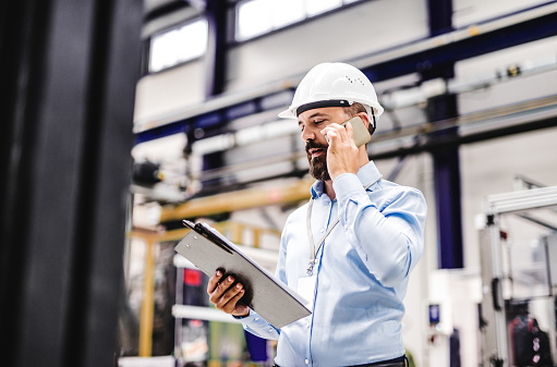 A portrait of a mature industrial man engineer with clipboard and smartphone in a factory, making a phone call. Copy space.