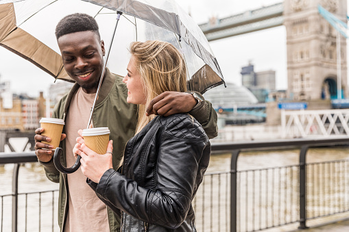 Happy multiracial couple walking in London holding an umbrella on a rainy day. Black man and white woman drinking tea or coffee, bonding and having fun together with Tower Bridge on background. Love and travel concepts